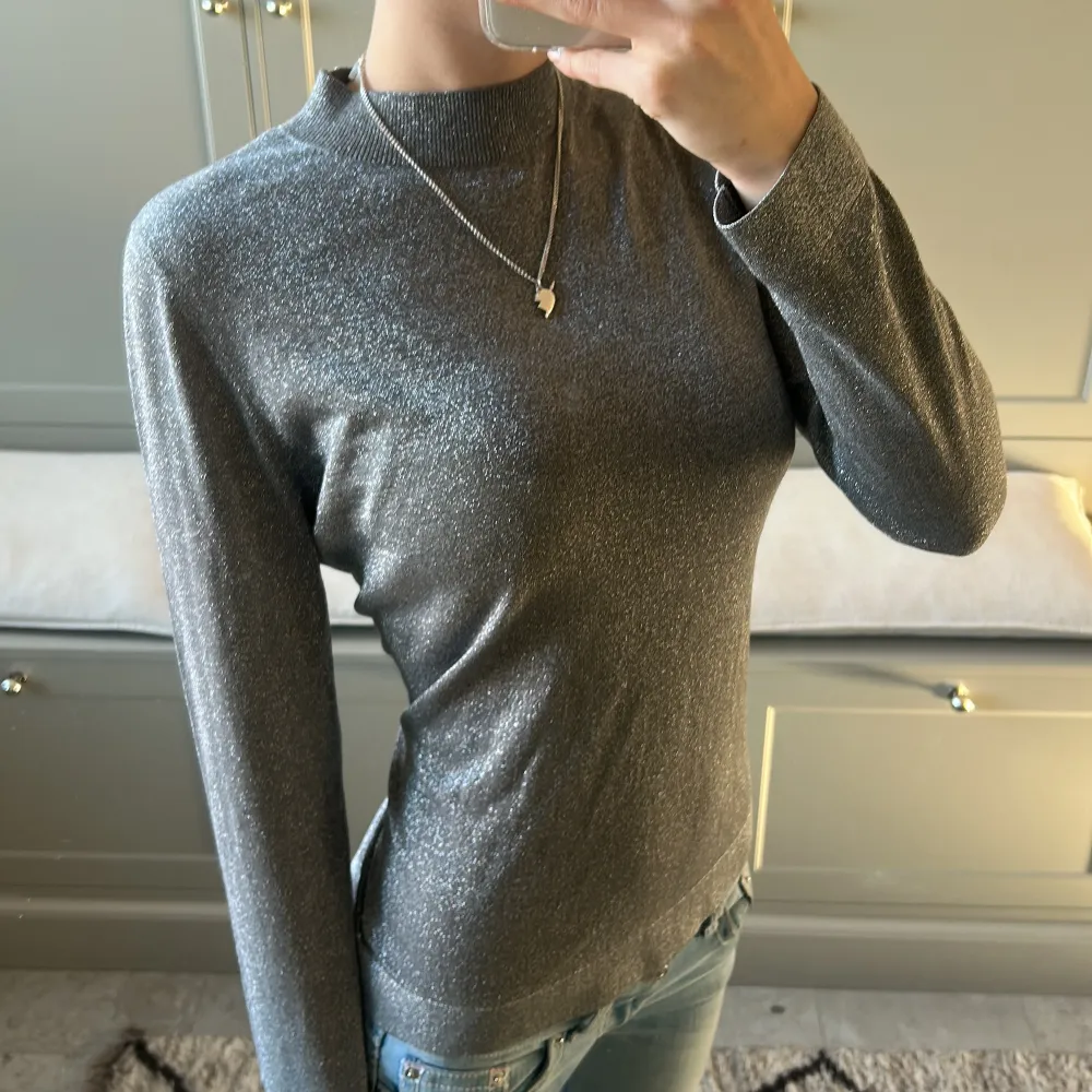 Snygg grå tröja med glittrig finish. Perfekt för att ge lite extra bling till din outfit. Tröjan har en hög krage och långa ärmar, vilket gör den både stilren och bekväm.. Neuletakit & Villapaidat.