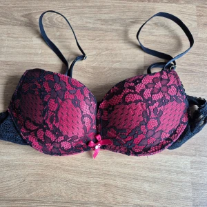 Röd och svart spets-bh storlek 70AA - Säljer en elegant röd bh med svart spetsdetalj från No Secret. Den har justerbara axelband och en liten rosett framtill. Lite lätt vadderad. Perfekt för att känna sig extra fin. Den har inga tecken på användning. Storlek 70AA / 32AA.