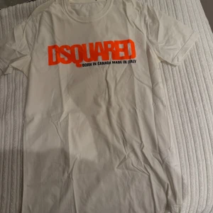 Vit t-shirt från Dsquared2 - Säljer en vit t-shirt från Dsquared2 med en iögonfallande orange logga på bröstet. Tillverkad i Italien med texten 'Born in Canada Made in Italy' under loggan. Perfekt för en stilren och modern look. Nexktag har fallit av , äkta dsquared2! 