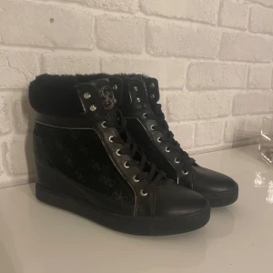  Klack sko från Guess - Snygga svarta sneakers från Guess med pälsdetalj runt ankeln och snörning framtill. Skorna har en diskret logga på sidan och en metallplatta med Guess-logga på hälen. Perfekta för en stilren look.
