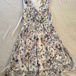 Blommig klänning från Vila - Säljer en vacker blommig klänning från Vila. Den är ärmlös med omlott-design och har ett fint blommönster i olika färger på en ljus bakgrund. Perfekt för sommarens alla tillfällen!