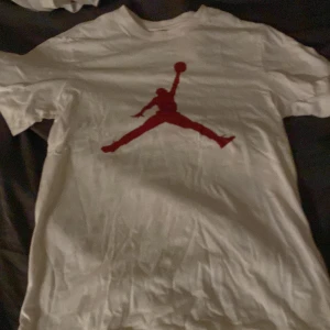 Vit t-shirt från Jordan - Säljer en vit t-shirt från Jordan med det ikoniska röda logotyptrycket på framsidan. Perfekt för sportiga tillfällen eller som en stilren vardagslook. T-shirten är kortärmad och gjord i mjukt bomullsmaterial.