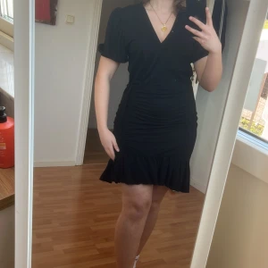 Svart kortklänning med volangdetaljer - Snygg svart kortklänning med v-ringning och volangdetaljer vid fållen. Klänningen har korta ärmar och en åtsittande passform med knytning i midjan för en smickrande silhuett. Dock är den bara som detalj och inte så att den knyter ihop hela klänningen. Den är en L men passar nog en M!