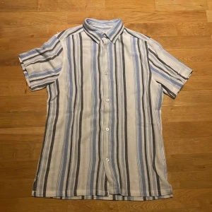 Randig skjorta från Smog - Säljer en stilren randig skjorta från Smog i blått, vitt och svart. Skjortan har korta ärmar och knappar framtill. Perfekt för en avslappnad look.