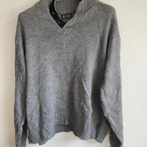 Grå stickad tröja från Chaps - Säljer en stilren grå stickad tröja från Chaps med ribbad krage och knappar vid halsen. Tröjan har långa ärmar och ribbade muddar vid ärmslut och nederkant. Perfekt för kyliga dagar!