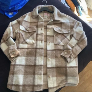 Beige och vit rutig overshirt - Säljer en snygg beige och vit rutig overshirt med knappar framtill. Jackan har två stora bröstfickor med lock och långa ärmar med knappdetaljer vid ärmsluten. Perfekt för lager-på-lager-stil!