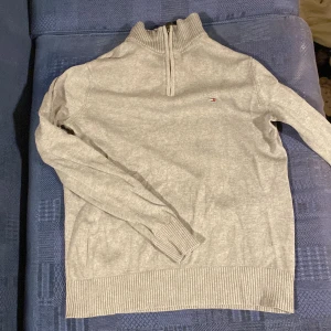 Grå tröja med dragkedja från Tommy Hilfiger - Säljer en stilren grå tröja från Tommy Hilfiger med dragkedja vid halsen. Tröjan har långa ärmar och ribbade muddar vid ärmslut och nederkant. Perfekt för en avslappnad look.