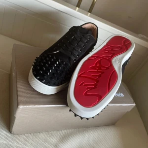 Svarta sneakers med nitar från Christian Louboutin - Snygga svarta sneakers från Christian Louboutin med nitar över hela ovandelen och den ikoniska röda sulan. Skorna har snörning och en vit sula som kontrasterar mot det svarta. Perfekta för att ge en edgy touch till din stil. Skriv för mer info eller prisdiskutering