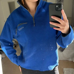 Blå fleece tröja från Everest - Säljer en blå fleece tröja från Everest med halv dragkedja och broderad logga på bröstet. Tröjan har en normal passform och långa ärmar, perfekt för kyliga dagar. Den har även en praktisk bröstficka med dragkedja.