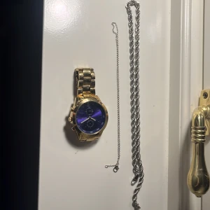 Guldfärgad klocka med blå urtavla - Snygg guldfärgad klocka med en djupblå urtavla och metallarmband. Perfekt för att ge en touch av elegans till din stil. Klockan har flera små urtavlor och en robust design.