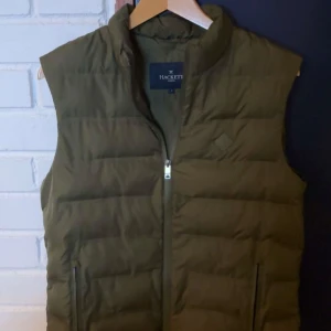 Olivgrön dunväst från Hackett - Snygg olivgrön dunväst från Hackett med dragkedja framtill. Perfekt för lager-på-lager under kyliga dagar. Västen har en stilren design med quiltade detaljer och en hög krage för extra värme.
