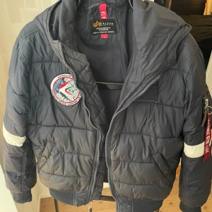 Mörkblå NASA bomberjacka - Säljer en snygg mörkblå bomberjacka från Alpha Industries med NASA-tryck på ryggen och Apollo-patch på bröstet. Jackan har dragkedja och ribbade muddar. Perfekt för dig som gillar rymdtema! 🚀