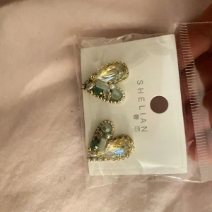 Glittriga örhängen från Shelian - Snygga glittriga örhängen från Shelian med en mix av gröna och gula stenar. Perfekta för att ge en extra touch till din outfit. De har en unik design med en kombination av olika former och färger.