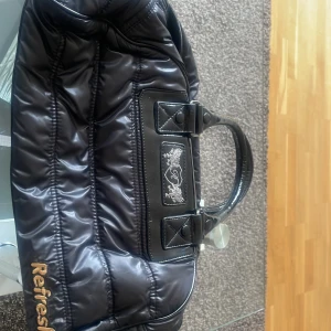 Svart axelväska från Refresh - Snygg svart axelväska från Refresh med quiltad design och glansigt material. Väskan har två handtag och en dragkedja upptill. Perfekt för att bära med sig det nödvändigaste med stil.