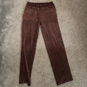 Bruna byxor från Juicy Couture - Snygga bruna byxor från Juicy Couture i mjukt material med broderad logga. Byxorna har en bekväm midja med snörning och praktiska fickor både fram och bak. 