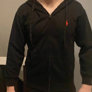 Svart hoodie från Ralph Lauren - Säljer en stilren svart hoodie från Ralph Lauren med dragkedja och det klassiska röda logotypen på bröstet. Perfekt för en avslappnad stil. Den har långa ärmar och en bekväm passform.