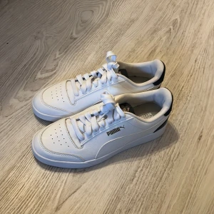 Vita sneakers från Puma - Snygga vita sneakers från Puma med svarta detaljer. Skorna har klassisk snörning och en stilren design med perforeringar på ovansidan för extra ventilation. Puma-loggan syns tydligt på sidan och hälen. Perfekta för en avslappnad och sportig look.Storlek 39 tyvärr ingen låda.Dem är i helt sprillans byt skick .ny pris 500kr
