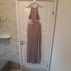 Ljuslila långklänning med satinband - Elegant ljuslila långklänning med plisserad design och vackra satinband som korsas i ryggen. Klänningen har en ärmlös stil och ett knytband i midjan för en smickrande passform. Perfekt för speciella tillfällen.