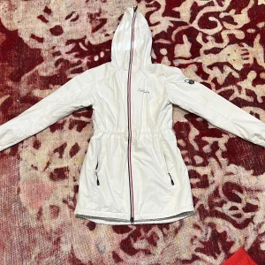 Vit vindjacka från Sail Racing - Snygg vit vindjacka från Sail Racing med huva och dragkedja. Jackan har en justerbar midja och två praktiska fickor med dragkedja. Perfekt för blåsiga dagar!