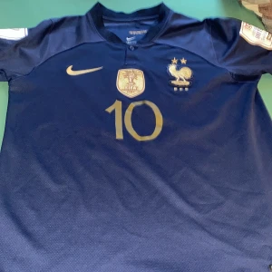Mbappe vm 2022 tröja  - Säljer en blå fotbollströja från Nike med guldiga detaljer. Tröjan har korta ärmar och är prydd med nummer 10 och namnet Mbappe på ryggen. Perfekt för fotbollsfans som vill visa sitt stöd för laget. Mbappe prestera jättebra med denna tröja vem vet kanske du gör samma om du köper den 😉