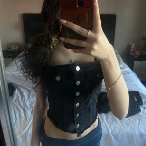 Svart jeanskorsett med knappar - Snygg svart korsett i jeansmaterial med silverfärgade knappar framtill. Den har en cool, rå kant och dekorativa fickor. Perfekt för att ge en edgy touch till din outfit!