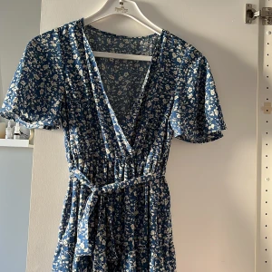 Blå blommig långklänning - Säljer en vacker blå långklänning med blommigt mönster. Klänningen har en v-ringad design och korta ärmar. Den kommer med ett knytbälte i midjan för en smickrande passform.