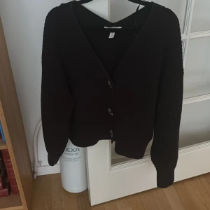 Svart kofta med knappar från H&M - En svart kofta med knappar framtill. Koftan har långa ärmar och en ribbad struktur. 