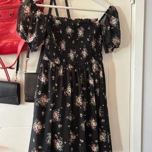Blommig klänning från Hollister - Säljer en söt blommig klänning från Hollister i storlek XS. Klänningen har en offshoulder-design med korta puffärmar. Den är svart med ett vackert blommönster i fina färger. Perfekt för sommarens alla tillfällen! 🌸