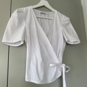 Vit omlottopp från NA-KD - Säljer en stilren vit omlottopp från NA-KD med korta ärmar och knytband i midjan. Perfekt för en chic och avslappnad look. Toppen har en v-ringad design som ger en elegant touch. Perfekt till fest, middag eller annat nu i vår och sommar!