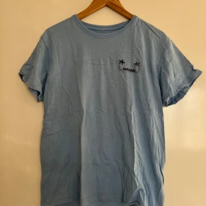 Blå t-shirt från Paradox Skateboards - Säljer en blå t-shirt från Paradox Skateboards med ett coolt tryck av en solnedgång och palm på baksidan. T-shirten har korta ärmar och en rund halsringning. Perfekt för skate-stil. Storlek M. Knappt använd.