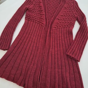 Vinröd stickad kofta av Alpacka & Ull blandning  - Säljer en vinröd stickad kofta med ett vackert mönster och långa ärmar. Koftan har en öppen design och är tillverkad i en blandning av Alpacka, ull och akryl. Perfekt för kyliga dagar. Lägg till ett guldfärgat bälte så blir det wow!