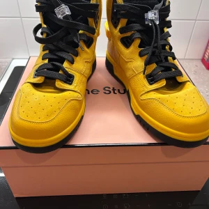 Gula sneakers med svarta detaljer - Snygga gula sneakers med svarta snören och detaljer. Skorna har en hög design med perforerade detaljer på ovansidan och en robust svart sula. Perfekta för en cool och avslappnad stil.