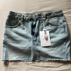 Ljusblå jeanskjol från Pull&Bear - Säljer en ljusblå jeanskjol från Pull&Bear med rå kant nedtill och klassiska fem fickor. Kjolen har dragkedja och knapp framtill samt bälteshällor. Perfekt för dig som gillar en chill och trendig denimlook.