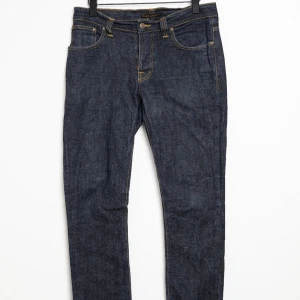 Mörkblå jeans från Nudie Jeans - Snygga mörkblå jeans från Nudie Jeans i klassisk femficksmodell modell "Grim Tim".Jeansen är tillverkade av 99% ekologisk bomull och 1% elastan för lite stretch. De har raka ben och kontrastsömmar i gult. Perfekta för dig som gillar stilrena och tidlösa jeans.
