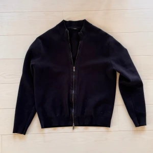 Svart stickad kofta med dragkedja från Massimo Dutti - Svart stickad kofta från Massimo Dutti med hel dragkedja framtill och ribbad krage. Tröjan har långärmade smala ärmar och är tillverkad i mjuk ull som känns skön mot huden. Perfekt för dig som gillar stilrena och enkla plagg med snygg passform. Storleken är L men passar lite mer som M