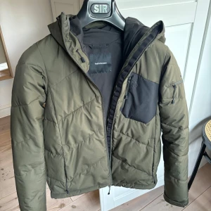 Olivgrön pufferjacka Peak Performance - Snygg olivgrön pufferjacka från Peak Performance med svart bröstficka och dragkedjor. Jackan har huva, flera praktiska fickor och coola detaljer som logga på ärmen. Perfekt för kalla dagar och skidåkning.