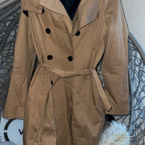 Beige trenchcoat med bälte - Snygg beige trenchcoat med klassisk dubbelknäppning och bälte i midjan. Jackan har långa ärmar, bred krage och två fickor framtill. Perfekt för dig som vill ha en stilren och tidlös look under vår och höst.