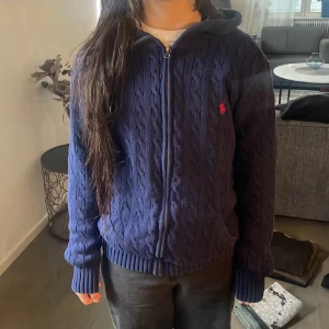 Mörkblå stickad hoodie från Polo Ralph Lauren - Säljer en mörkblå stickad hoodie från Polo Ralph Lauren med dragkedja och huva. Tröjan har kabelstickat mönster, långa ärmar och en röd Polo-logga broderad på bröstet. Perfekt för dig som gillar klassisk stil med en modern twist.