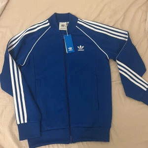 Blå Adidas track jacket med dragkedja - Snygg blå Adidas track jacket med klassiska vita ränder längs ärmarna och broderad logga på bröstet. Jackan har ribbade muddar, ståkrage och hel dragkedja framtill. Tillverkad i mjuk polyester som ger en sportig vibe. Perfekt för chill eller träning.