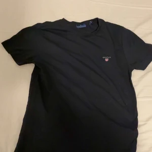 Svart t-shirt från GANT - Svart t-shirt från GANT med klassisk rund hals och korta ärmar. Diskret GANT-logga med sköld på bröstet. Enkel och stilren design som funkar till jeans eller shorts. Mjuk bomullskvalitet för skön känsla hela dagen.