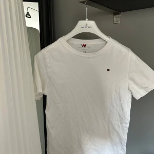 Vit t-shirt från Tommy Hilfiger - Säljer en klassisk vit t-shirt från Tommy Hilfiger med liten broderad logga på bröstet. T-shirten har rund hals och korta ärmar, perfekt för en clean och enkel stil. Materialet är mjuk bomull som känns skönt mot huden.