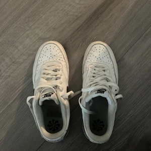 Nike Air Force 1 vita sneakers - Säljer ett par klassiska Nike Air Force 1 sneakers i vitt med svart swoosh på sidan. Skorna har perforerad tå, snörning och platt sula. Tillverkade i skinn med låg profil och rund tå. Perfekta för dig som gillar clean och tidlös streetstil.