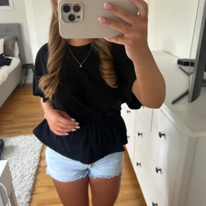 Svart blus med resår i midjan - Säljer en svart blus med korta ärmar och rund hals. Toppen har en markerad resår i midjan som ger en snygg volang-effekt nedtill. Perfekt för dig som gillar en enkel men trendig look. Materialet känns mjukt och lätt, passar bra till både jeans och kjol.