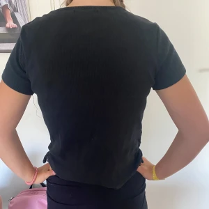 Svart ribbad t-shirt från XOXO Friends - Svart ribbad t-shirt från XOXO Friends i mjuk bomull med lite stretch. Modellen är kortärmad och har snörningsdetalj i sidan för en cool look. Perfekt till jeans eller shorts när du vill ha en enkel men snygg outfit.