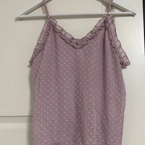 Ljuslila prickig linnetopp Bik Bok - Supersöt ljuslila linnetopp från Bik Bok med tunna axelband och volangdetaljer längs urringningen. Toppen har ett prickigt mönster och är gjord i ett lätt, glansigt material som känns luftigt och fräscht. Perfekt för att addera lite extra gullig vibe till din outfit.