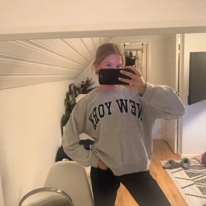 Grå sweatshirt med New York-tryck - Säljer en grå sweatshirt med stort mörkblått New York-tryck över bröstet. Tröjan har en loose passform, rund hals och långa ärmar. Perfekt för chill dagar och enkel att matcha med jeans eller leggings.