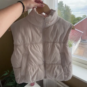 Beige dunväst från H&M - Säljer en beige dunväst från H&M i storlek S. Västen har hög krage, dragkedja framtill och två fickor. Den är quiltad och har en puffig, trendig look som passar perfekt till lager-på-lager-stil.