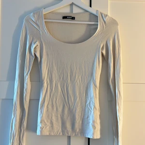 Vit långärmad topp från BikBok - En basic vit/ beige långärmad topp från BikBok med rundad, djup urringning. Enkel och stilren modell i mjuk bomull som passar perfekt till jeans eller kjol. Snygg att bära både som den är eller under en jacka.