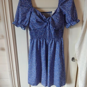 Blommig klänning - En blå kortklänning med små vita blommor över hela tyget, puffiga ärmar och resår midja. Klänningen har en fastsyd knytning framtill mellan bysten, dock går det att knyta om den till en rosett. Bröst kuporna har också vad i sig så att man kan ha klänningen utan bh. Den har även små volangdetaljer vid ärmarmas kant. Klänningen är tunn och luftig och kan användas en vanlig sommardag men också till en mer uppklädd tillställning. Lappen sitter fortfarande kvar.