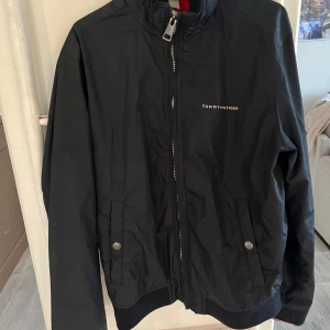 Marinblå vindjacka Tommy Hilfiger S - Svart vindjacka från Tommy Hilfiger i storlek S. Jackan har dragkedja framtill, två fickor med tryckknappar och ribbade muddar. Klassisk logga på bröstet och insidan har snygga Hilfiger-ränder. Perfekt för dig som gillar stilren och sportig look.
